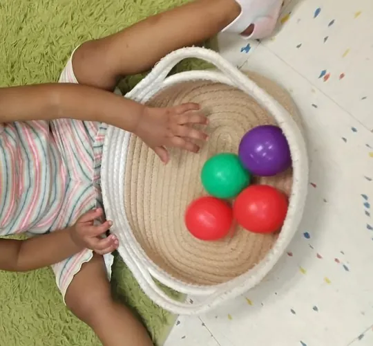 A toddler's hands filling a woven basket with red, green and purple balls.