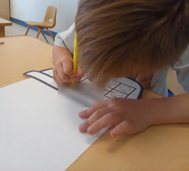 A pre-K child bends over a ten-frame worksheet with a yellow pencil.