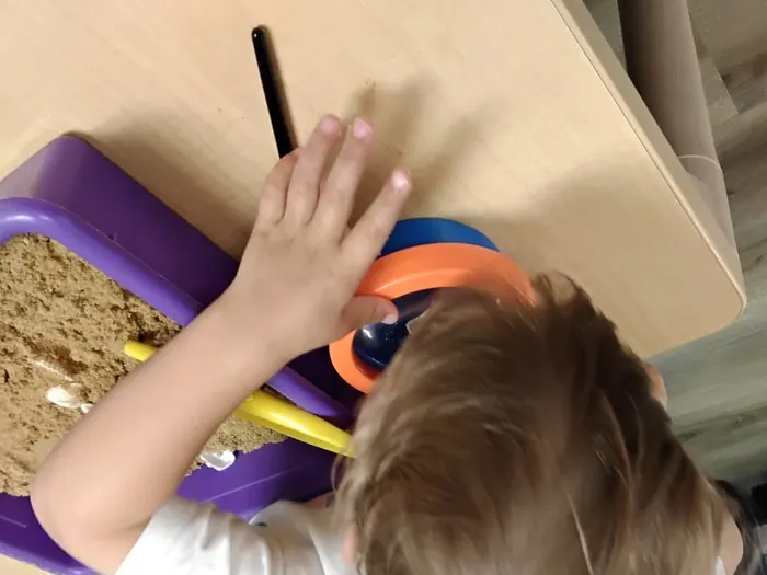 A two-year-old's hand reaches into a purple bin of sand with shells, seen from above.