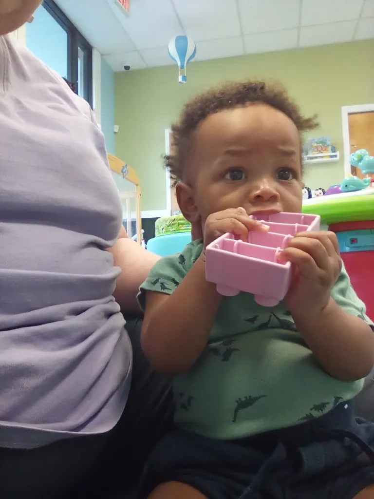 A baby holds a pink toy block, sitting with a teacher's support.