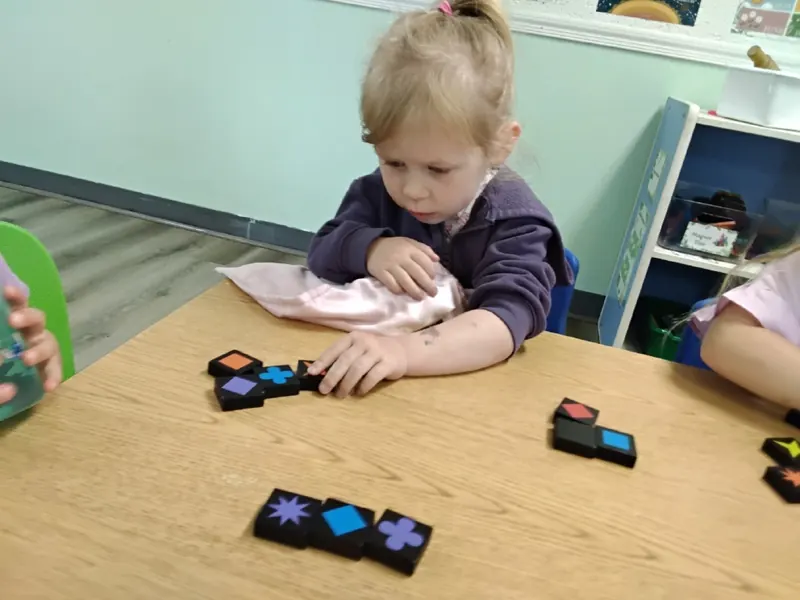 A preschooler arranges black tiles with colored shapes on a table.