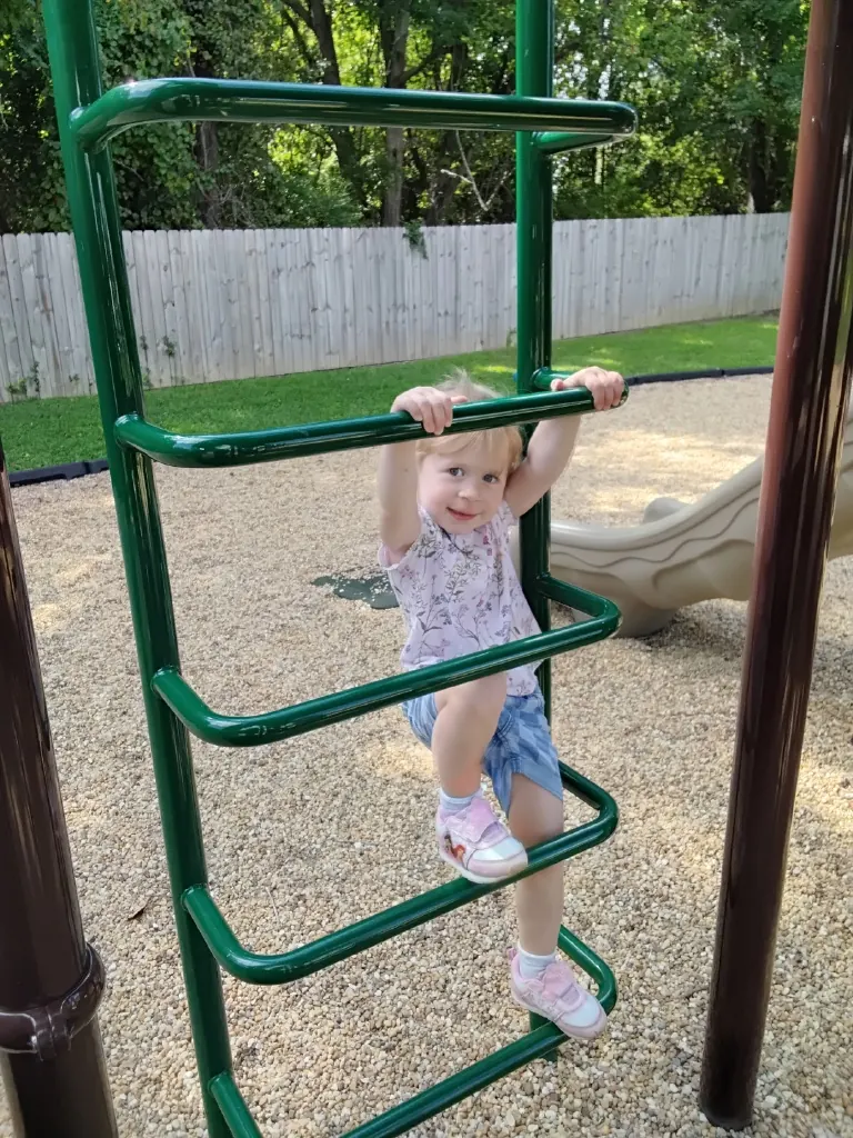 A preschooler climbs a green ladder on the playground.