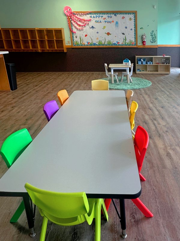 An ocean-themed Falmouth preschool classroom with a long table ringed by rainbow-colored chairs and a Happy to Sea You bulletin board.