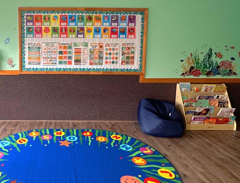 A Falmouth circle-time area with an under-the-sea calendar and learning board, a book nook with a bean bag, and a blue alphabet rug.
