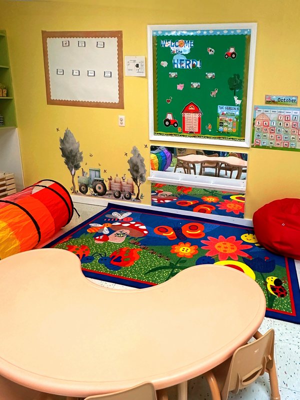 A farm-themed Falmouth toddler room with a Welcome to the Herd board, a garden play rug, a crawl tunnel, and a kidney-shaped table.
