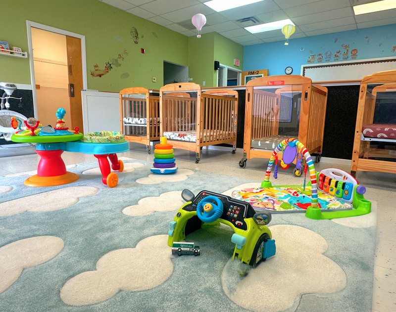 The Falmouth infant room with wooden cribs, soft activity centers, and a cloud-patterned rug.