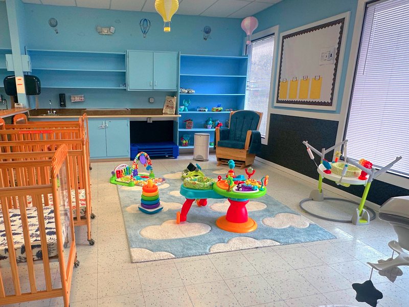 A bright Falmouth infant room in soft blue, with cribs, a glider rocker, and infant activity stations on a cloud rug.