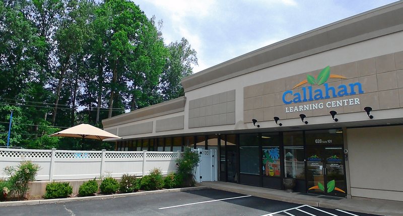 The sunny storefront entrance of Callahan Learning Center in Falmouth, with the Callahan logo above the door, a shaded patio table, and a white privacy fence.