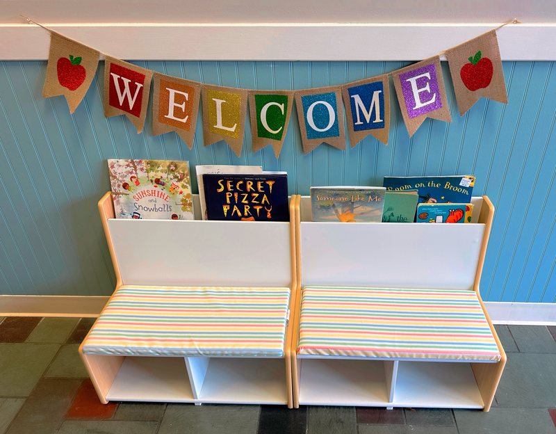 A welcoming reading nook in the Falmouth lobby, with a colorful WELCOME banner, a picture-book display, and cushioned benches.