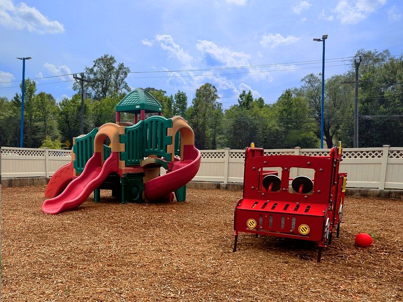 The fenced Falmouth playground under a blue sky, with a climber, slides, and a red fire-truck play structure.