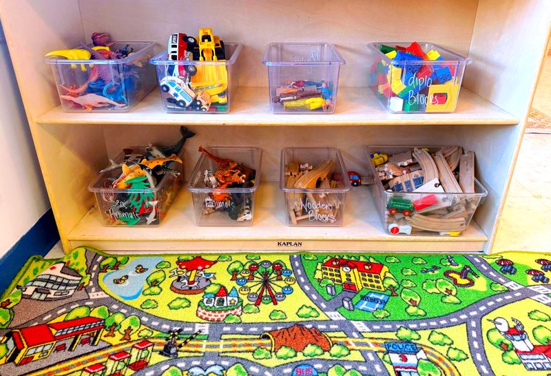 Labeled bins of animals, blocks, and wooden train tracks above a colorful road-and-town play rug in a Locust Grove classroom.