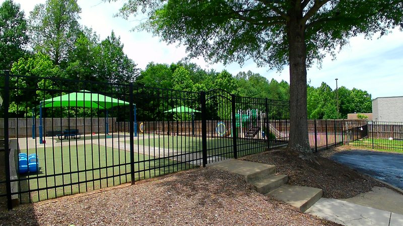 The Locust Grove playground with turf play areas under green shade canopies, enclosed by a black metal fence and framed by shade trees.