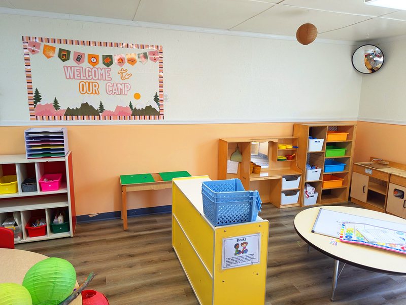 A Maple Grove classroom with a Welcome to Our Camp mountain-themed board, cubbies of colorful bins, and a rounded activity table.