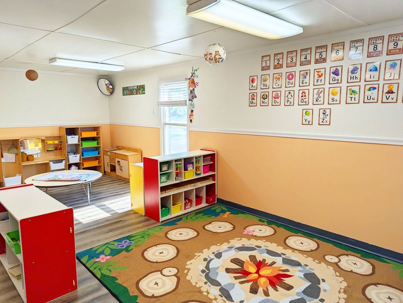 A Maple Grove classroom lined with alphabet and number charts, colorful bins, and a campfire-themed circle-time rug.