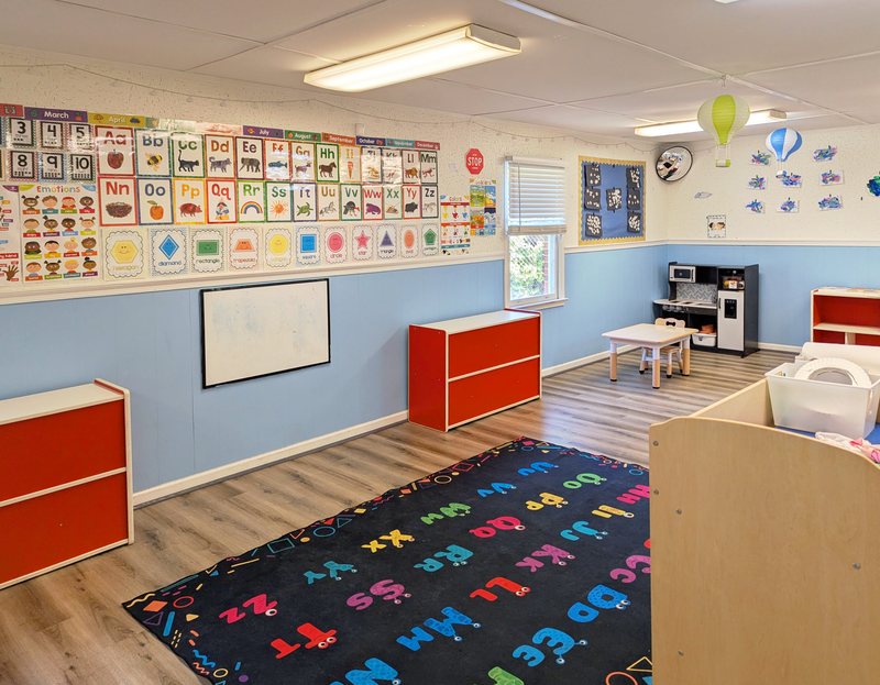 A Maple Grove preschool room with a full alphabet-and-calendar wall, an ABC play rug, a play kitchen, and red-accented shelves.