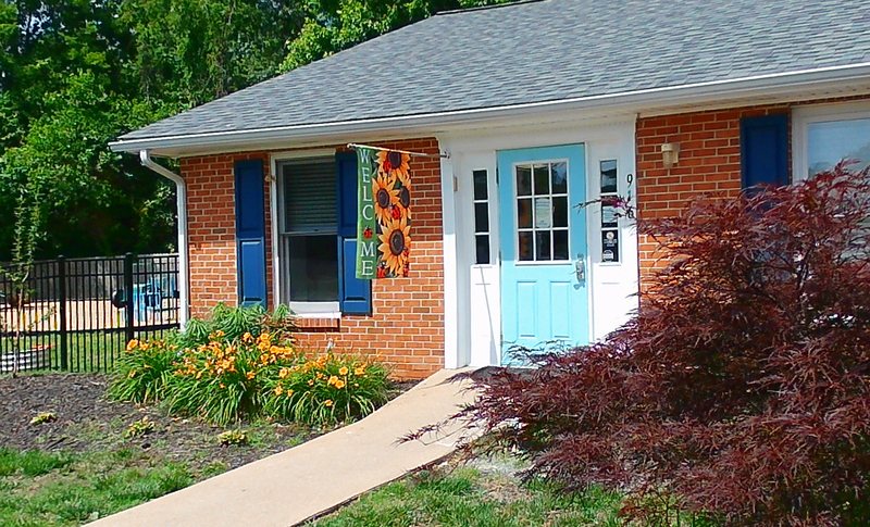 The Maple Grove center's brick entrance with a teal front door, a sunflower Welcome banner, blue shutters, and a blooming daylily garden.