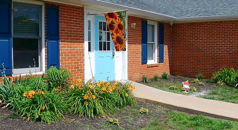 A wider view of the Maple Grove brick building and teal entry door, framed by flower beds and a friendly garden gnome.
