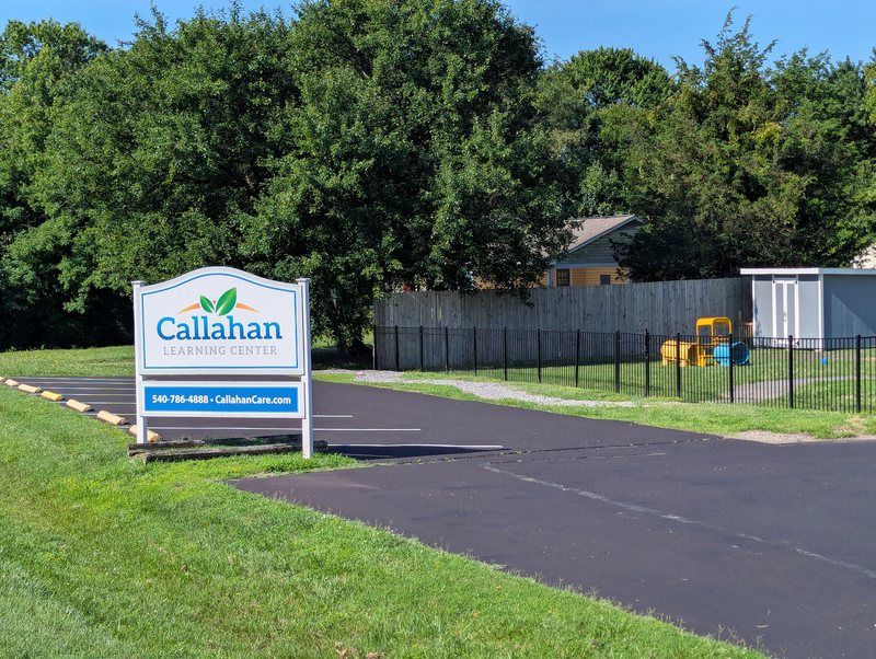 The Callahan Learning Center sign at the Maple Grove entrance drive, with the fenced playground and building beyond.