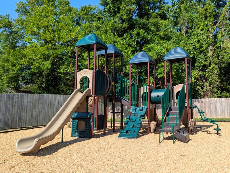The Maple Grove playground with a large climbing structure and slide on soft mulch, surrounded by leafy trees.